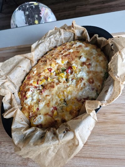 A baked, oval-shaped dish, likely a casserole or quiche, is presented in a dark, round baking pan lined with crumpled, browned parchment paper. The dish features a golden-brown, melted cheese crust with visible yellow corn kernels, red bell pepper fragments, and white cheese crumbles. The pan rests on a light-colored wooden surface with visible grain. In the blurred background, a portion of a spherical object displaying black, white, and pastel-colored patterns, including stars, is partially visible against a light-colored vertical plane. No human subjects are present. The scene is located in Niedernhausen, Germany.