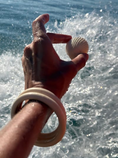 A close-up, vertical shot shows a hand holding a small, white, ridged seashell against a background of choppy blue water with sunlit reflections. The hand is held up, with the thumb and forefinger pinching the shell. A thick, cream-colored bangle is visible on the wrist. The water is blurred and appears to be in motion, creating a dynamic backdrop. The scene suggests an outdoor, possibly coastal, setting, consistent with locations like Baan Thong Krut, Thailand, known for its beaches and marine life.