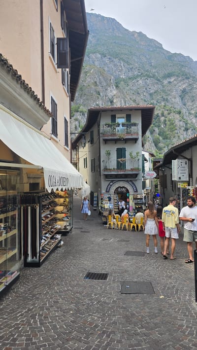 This image captures a street scene in Limone sul Garda, Italy, oriented vertically. The backdrop is dominated by a steep, rocky mountain covered in sparse vegetation. In the foreground, a cobblestone street lines the right side of the image, with several people walking and standing.

On the left side, buildings rise up, displaying architectural details like windows with shutters and balconies with plants. An awning extends from one building, partially covering a shop entrance. The shop appears to sell clothing or accessories, with items displayed behind glass.

Visible text on the awning reads "OLGA e ROBERTO". Above this, a sign for "BAR CAFFÈ GELATERIA" is prominent on another building, featuring a circular logo with the word "PINK PANTHER" above it. Another sign further up the street reads "LOTTO - TABACCHI - SERVIZI".

Several individuals are depicted on the street, some casually dressed for warm weather, suggesting it is daytime. One group is gathered near the bar, while others are walking along the street. The overall atmosphere suggests a leisurely tourist day. The lighting indicates it is likely late afternoon or early evening.