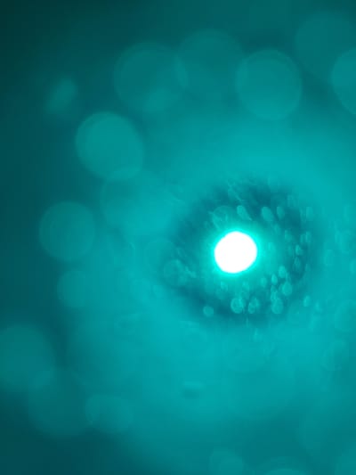 The image is an abstract, out-of-focus shot with a teal-colored background. The primary subject is a bright, white, circular light source in the center of the frame, creating a bokeh effect. Surrounding this central light are numerous smaller, blurred circular shapes, also in varying shades of teal and light green. These appear to be reflections or light sources that are not in sharp focus. The overall impression is one of softness and diffusion, with the bright light in the center drawing the viewer's eye. There is no discernible scene, objects, people, or text. The time of day, weather, and location cues are not apparent due to the abstract nature of the image. The emotional tone is somewhat ethereal and calming due to the soft lighting and color palette.