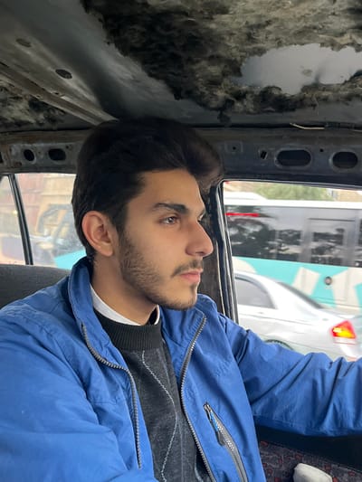 The image displays a young male seated inside a vehicle, positioned in the Al-Qaboun Municipality, Syria. The subject is shown from a side profile, looking towards the right. He has dark hair styled with significant volume, dark eyebrows, and a light beard and mustache. He is dressed in a white collared shirt, a dark patterned sweater, and a blue zip-up jacket with a chest pocket.

The interior of the vehicle exhibits signs of considerable wear; specifically, the headliner is severely damaged and detached in multiple areas, exposing the vehicle's structural frame and dark, fibrous insulation material.

Through the vehicle's side window, a blurred urban street scene is visible. Moving traffic includes a large, dark-colored bus featuring a light blue and white abstract pattern on its side, followed by a white sedan with illuminated red tail lights. The background shows indistinct shapes, possibly buildings, further indicating a city setting. The male's visible right arm rests within the car's interior. No overt actions or interactions are depicted.