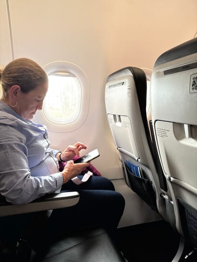 The scene is set within a commercial aircraft cabin, presumably in transit to or from the Area Metropolitana Centro Occidente (AMCO), Colombia. The specific ground location within AMCO is not discernible.
An adult female, appearing middle-aged, with light brown hair pulled back, occupies a window seat on the aircraft's left side. She wears a light blue collared button-up shirt, dark trousers, a silver hoop earring in her left ear, and a dark beaded bracelet on her left wrist. Her gaze is directed downwards at a dark-colored smartphone, which she holds horizontally with both hands, actively interacting with its touchscreen with her left index finger. A purple or magenta fabric item is draped across her lap, partially covering a fastened seatbelt.
To her right, the seat directly in front, an unoccupied passenger seat, features a black upholstered back and a light gray plastic shell. This shell includes a fold-down tray table mechanism, a safety card holder, and a mesh pocket containing blue-covered documents. A QR code and the "Avianca" brand name are visible on the upper portion of this seat-back. The aircraft interior exhibits light beige overhead panels and a circular window adjacent to the seated female, through which bright ambient light and indistinct exterior features are visible. The flooring is dark-colored.