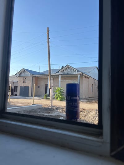 The image, taken from inside a room looking out through a window, shows a residential street scene in Hazorasp, Uzbekistan. The focus is on a row of houses with light-colored stucco walls and dark metal roofs. One house features a distinctive ornamental detail above its entrance and a large, dark garage door. Another house, partially visible to the left, has a brick facade and a dark-tiled roof. A wooden utility pole with multiple wires stands prominently in the middle ground. The street appears to be unpaved, with gravel and dirt visible. In the foreground, a blue cylindrical object, possibly a glue stick or aerosol can with the brand "VIBRO" and the text "VB 012" visible, is placed on the windowsill, partially obscuring the view of the street. The sky is clear and blue.