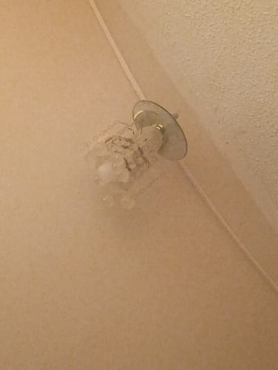 The media file displays a close-up, angled shot of a ceiling with a textured finish, exhibiting small yellow flecks against a pale beige background. A white trim runs diagonally across the upper portion of the frame. In the lower half of the image, a broken light fixture is visible, hanging from the ceiling. The fixture appears to be made of metal and glass, with shards of glass and wire hanging down, suggesting damage. A thin, white cord also dangles from the fixture. The lighting in the image is dim, and there are no discernible people or other objects. The scene is indoors, likely a residential or commercial space, given the ceiling and light fixture. There is no visible text, and the overall impression is one of disrepair or neglect. The location context of Puerto La Cruz, Venezuela, provides a geographical reference but does not offer specific visual cues within the image itself.