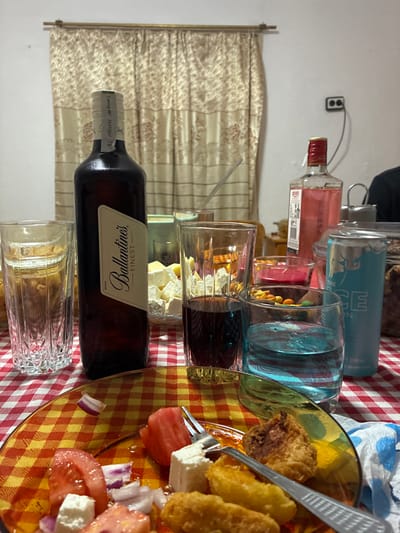 The scene depicts an indoor domestic environment, specifically a dining table in Plodovitovo, Bulgaria. The table is covered with a red and white checkered tablecloth.
In the foreground, a clear glass plate with a yellow and red checkered pattern holds remnants of a meal. Visible items on the plate include several slices of red tomato, cubes of white feta cheese, slices of red onion, and multiple golden-brown pieces of fried food. A silver fork rests on the plate, partially covered by food items. A crumpled blue and white paper napkin is positioned to the lower right of the plate.
Moving towards the mid-ground, a tall, dark brown glass bottle of "Ballantine's Finest" blended Scotch whisky stands upright, appearing approximately three-quarters full. To its left, a tall, clear, patterned glass is mostly empty. To the right of the whisky bottle, another tall, clear, patterned glass contains a dark red liquid, about one-third full. Further right, a wide, clear glass holds a bright blue liquid, roughly half full. A light blue energy drink can, labeled "THE EDGE," stands behind this glass. Behind the blue liquid glass, a small, dark red candle is visible in a clear holder, positioned in front of a small bowl containing assorted colored snacks. A clear glass bowl containing numerous white feta cheese cubes is situated behind the red liquid glass.
In the background on the table, a clear glass bottle with a red cap contains a pink liquid. A silver metallic teapot is visible further to the right. A long, thin, clear glass rod or straw rests horizontally above the mid-ground items. The background wall is white, featuring a window covered by a light-colored curtain with a patterned design, hung on a gold-colored rod. A white electrical outlet with two circular sockets is visible on the upper right portion of the wall. A dark-clad shoulder and head of an individual are partially visible on the far right, suggesting at least one person is seated at the table. The overall scene suggests a gathering or meal in progress or recently concluded.
