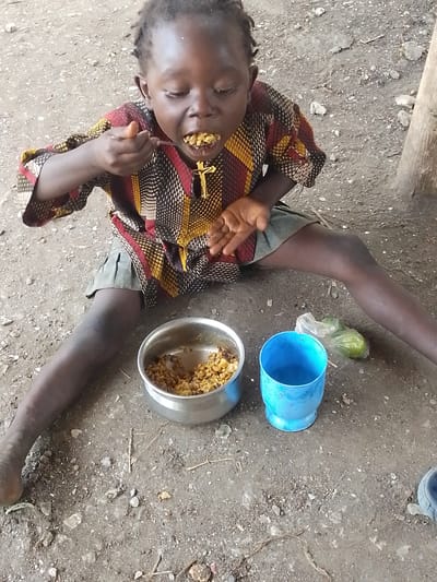 A young child is sitting on the ground in what appears to be an outdoor, unpaved area, likely in Otulu, Nigeria. The child is wearing a colorful, patterned shirt and a dark skirt. They are in the process of eating from a metal bowl with a spoon, bringing food to their mouth. A blue plastic cup and a small plastic bag containing some green items are placed on the ground near the bowl. The ground is covered with dirt, small stones, and some dried plant debris. A wooden pole is visible on the right side of the image. The child's expression suggests focus on eating. The lighting implies it is daytime.