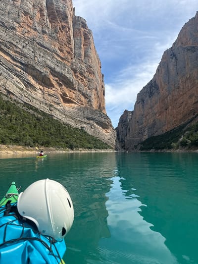 The image captures a first-person perspective from a kayak navigating the Congost de Mont-rebei gorge, located near Àger, Spain. The foreground features the bow of a green kayak, on which a blue dry bag secured by a black bungee net holds a white, water-splashed helmet. The kayak floats on a tranquil, highly saturated turquoise body of water, which exhibits subtle ripples and reflections of the surrounding rock formations and sky.

To the left and right, immense, vertical limestone cliffs with layered striations of grey and reddish-orange dominate the scene, indicative of a deep canyon. The cliff faces show varied textures and some sparse green vegetation at their bases and on ledges. In the mid-ground, another individual in a bright lime green kayak is visible, paddling with a yellow oar, moving away from the viewer towards the narrowing passage of the gorge. The kayaker is wearing a red life vest. Further into the distance, the canyon walls appear to converge, with a distant path or ledge visible along the lower section of the right-hand cliff. The sky above is partially cloudy, with scattered white clouds against a pale blue backdrop.