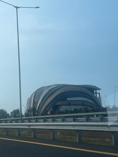 The image captures a daytime view of the Indonesia Convention Exhibition (ICE) building in Kosambi Barat, Indonesia, as seen from a moving vehicle on an elevated highway. The architectural structure of ICE is prominently featured, characterized by its distinctive curved, segmented roof resembling an open shell or a ribbed dome. The building's facade appears to be a mix of glass and darker, curved panels.

In the foreground, a portion of the asphalt highway is visible, marked with a bright yellow line. A metallic highway barrier separates the road from the area behind it, where the ICE building stands. Beyond the barrier, there is a strip of green grass and some vegetation, including palm trees, suggesting a tropical environment. A tall, slender streetlamp with a single light fixture stands to the left of the ICE building, extending vertically against a clear, pale blue sky. The overall lighting indicates it is daytime with good visibility. The perspective suggests the photograph was taken from a car window, looking towards the right. There are no people visible in the frame.
