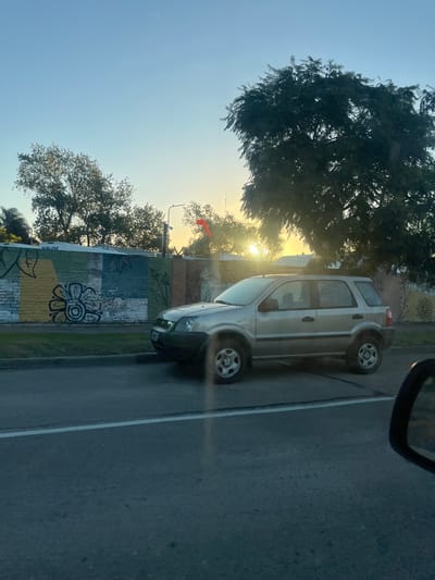 A silver SUV is driving on a street in Sarandí, Argentina, next to a mural painted on a brick wall, with trees and the setting sun visible in the background. The SUV is positioned between the dividing lines of the roadway. A rearview mirror from the vehicle taking the photograph is visible in the upper right corner.
