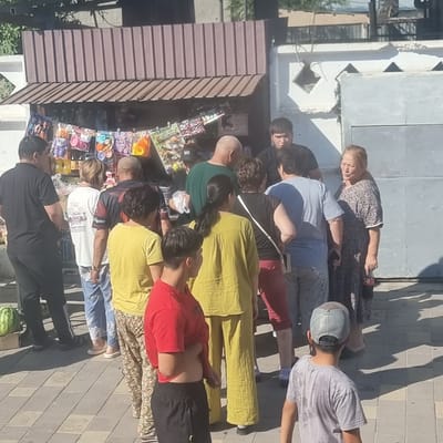 The image depicts an outdoor scene in front of a small, open-air stall in Turkistan, Kazakhstan. A group of people, appearing to be locals, are gathered around the stall. The stall displays a variety of small items, possibly toys or snacks, hanging from a rack. The people are dressed in casual clothing suitable for warm weather. Some individuals are interacting with the stall, while others are observing. The background shows a simple building structure with a corrugated metal roof and a white decorative wall. The ground is paved with interlocking bricks. The lighting suggests it is daytime, with visible shadows cast on the ground.