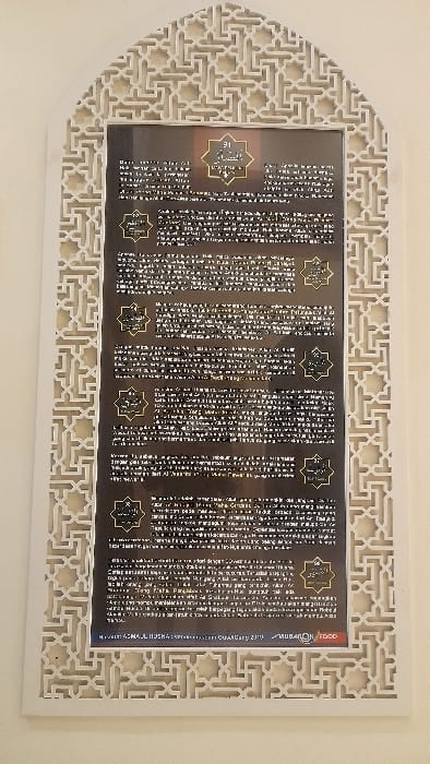 A vertical, arch-topped display board is centrally mounted on a smooth, light-colored wall. The board is encased in a light-toned, ornate frame exhibiting intricate geometric Islamic patterns, including interlocking stars and lines. The display panel features a dark background with multiple sections of text, primarily in Indonesian, each section initiated by a gold-colored, star-shaped emblem containing Arabic script. At the bottom left of the display, the text "MASJID ASMAUL HUSNA WWW.MASJIDASNAULHUSNA.COM Glantengan 2019" is visible, alongside a "MUBAROK FOOD" logo on the bottom right. The scene is static, devoid of people, subjects, actions, or interactions. The location is inferred to be within or adjacent to Masjid Asmaul Husna in Glantengan, Indonesia.