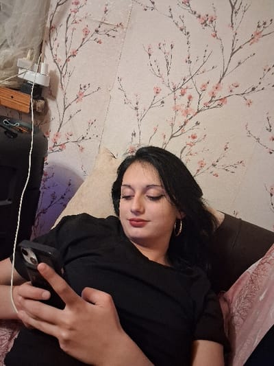 An indoor scene within the city of Lanchkhuti, Georgia, depicts a young adult female reclining, oriented to her left, looking downwards at a dark-colored smartphone held in her left hand. Her right thumb is positioned over the phone's screen. She has dark hair, dark eyeliner, and red lipstick, wearing gold-colored hoop earrings and a black short-sleeved top. The subject is resting against a light beige pillow, with a pink patterned fabric partially visible in the lower right foreground. The background wall is covered with wallpaper featuring pink cherry blossoms on dark branches against a light background. Visible vertical seams and minor edge separation are present in the wallpaper. In the upper left quadrant of the image, a white multi-outlet power adapter is plugged into a wall outlet, with white cables extending from it, positioned above a partial view of a wooden block or shelf fixture.