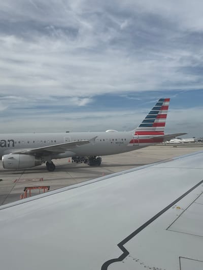 An American Airlines plane is parked on the tarmac under a cloudy sky. Its tail, adorned with red, white, and blue stripes, is prominent. The plane's registration number, N913US, is visible on its fuselage. The wing of another aircraft occupies a large portion of the right side of the frame, suggesting the photo was taken from a passenger window. Ground support equipment is stationed near the parked plane.