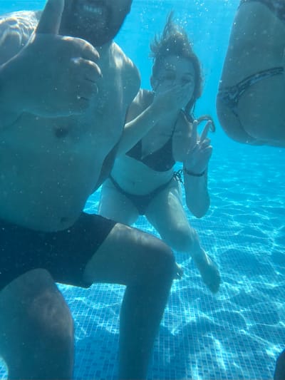 This underwater photograph captures a group of people enjoying a swimming pool. The image is dominated by the blue hues of the water and the tiled bottom of the pool, which is illuminated by sunlight creating dappled patterns. Several individuals are visible, mostly from the torso down. One person, likely a woman, is in the center of the frame, wearing a black bikini top and appearing to be submerged and possibly interacting with someone. Her face is partially obscured by her hand. Another person's legs are visible on the left, wearing dark swimming trunks. On the right, another person's buttocks and lower body are visible, wearing a patterned bikini bottom. The overall impression is one of casual recreation and enjoyment in a swimming pool setting. No specific time of day, weather conditions, or emotions are explicitly discernible, other than the general context of leisure. There is no visible text in the image. The setting is identified as Chiflik, Bulgaria.