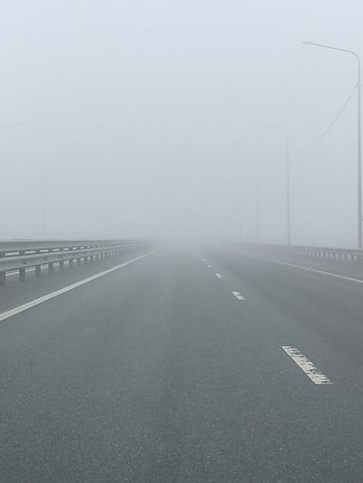 The image shows a highway shrouded in heavy fog. The location is identified as Сапуголи, Russia. The road surface is asphalt, marked with white lane lines and dashed center lines. Guardrails line both sides of the highway. Streetlights are visible, obscured by the fog. The overall visibility is severely limited due to the dense fog, reducing the perception of distance and depth. No people or vehicles are visible.