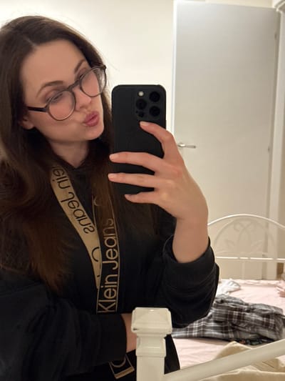 A female individual is depicted in a mirror selfie taken indoors in Scafati, Italy. She has long, dark brown hair and wears dark, round-framed eyeglasses. Her lips are pursed into a pouting expression. She holds a black smartphone with a multi-lens camera array in her right hand, angled to capture the reflection. She is dressed in a dark, long-sleeved top, with a beige strap featuring the text "Klein Jeans" visible diagonally across her chest. The reflected background includes a white door with a metallic handle to the upper right quadrant of the image. A white metal headboard or footboard of a bed is visible in the lower right, with folded plaid fabric resting on the bed.