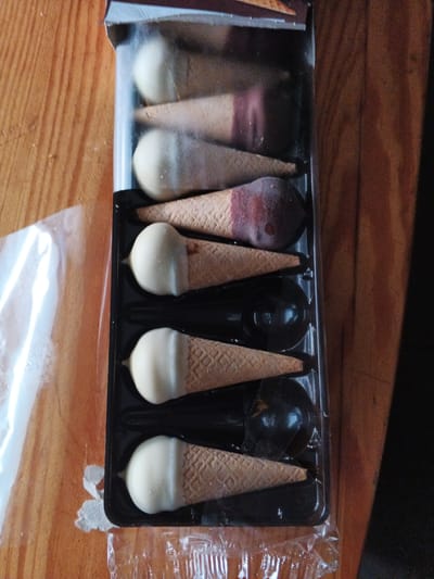 A black plastic tray filled with miniature ice cream cone-shaped chocolates sits on a wooden surface. The chocolates are arranged in two vertical columns of three. The cones are a light brown, waffle-like material. The tops of the cones are dipped in various coatings. From top to bottom, the first chocolate has a white coating, the second has a dark chocolate coating with some pinkish-red streaks, the third is entirely white, the fourth is entirely dark chocolate, the fifth is white, and the sixth is entirely dark chocolate. The tray is partially covered by a clear plastic wrap, which is slightly crinkled. The wooden surface appears to be a tabletop or countertop, with visible grain and a warm, light brown color. The scene is brightly lit, suggesting it might be daytime. There is no visible text in the image. The setting appears to be indoors, possibly a kitchen or dining area in Brzeg, Poland. The overall impression is a close-up product shot of confectionery.
