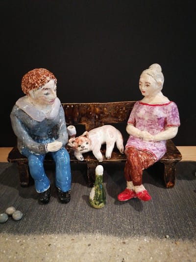 This is a close-up, eye-level, full-shot photograph of a ceramic diorama featuring two figures and a cat seated on a bench. The diorama is set against a plain black background. On the left, a figure with short, reddish-brown hair, wearing a blue long-sleeved shirt with a light blue collar and blue pants, sits with their hands clasped. To the right of this figure, a small, white cat with pinkish-orange markings lies down. Adjacent to the cat, on the bench, is a small green bottle with a white top. On the right side of the bench sits a figure with their hair in a bun, wearing a light purple dress with red trim and red leggings. This figure has their hands clasped in their lap. The bench itself is dark brown and appears to be made of textured ceramic. In the foreground, on the left, are three small, grey, pebble-like objects. The ground beneath the bench is textured and grey, with a sandy area in the very bottom of the frame. The overall impression is a staged scene, likely indoors given the controlled lighting and black background, with no discernible connection to Grünwald, Germany.