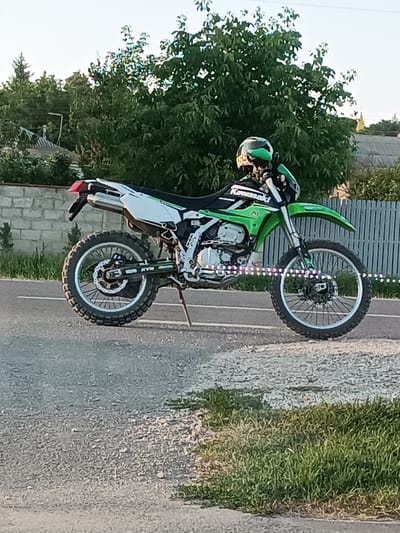 The image features a green and white Kawasaki dirt bike parked on a paved road. A person wearing a green and black helmet is seated on the bike, appearing to be taking a break. The bike has knobby tires, suggesting it is intended for off-road use. There's a string of small, colorful lights wrapped around the front wheel spokes, adding a decorative element. In the background, there's a grey brick wall, a light-colored wooden fence, and lush green trees. The setting appears to be a rural area, possibly in Grigorăuca, Moldova, with houses and other vegetation visible behind the fence. The lighting suggests it is daytime, likely late afternoon or early evening, given the soft light and shadows. The overall mood seems relaxed and casual. The word "Kawasaki" is visible on the side of the motorcycle.
