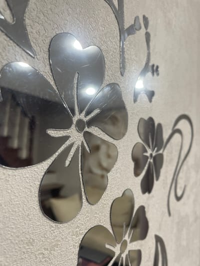 The image shows a close-up view of decorative mirrored floral decals affixed to a textured, light-colored wall. The decals are silver and reflect the surrounding environment, including what appear to be furniture and possibly lights. The floral designs are stylized, with distinct petal shapes and central circular elements. The texture of the wall is rough, resembling a stucco or heavily textured wallpaper. The lighting in the scene causes bright highlights and some glare on the mirrored surfaces, obscuring some details. There are no people or explicit actions depicted. The location is noted as Aropakkuzi, Russia, though the image itself does not provide visual cues to confirm this specific geographical context beyond what is provided in the prompt.