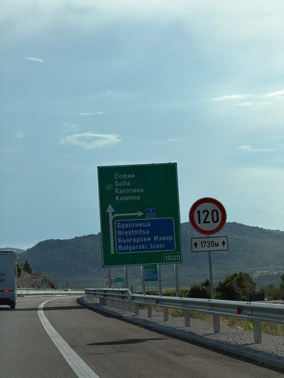 The image shows a road scene with a green directional sign indicating Sofia and Kalotina, as well as Brestnitsa and Bulgarski Izvor. A circular speed limit sign displays "120" and a supplementary sign indicates "1730m". The road curves to the right, with a metal guardrail along the side. In the background, a lush, tree-covered mountainside is visible under a partly cloudy sky. The location is identified as Peshtera, Bulgaria, however, there is no visual information in the image to confirm this specific location. The scene appears to be on a highway or major road.