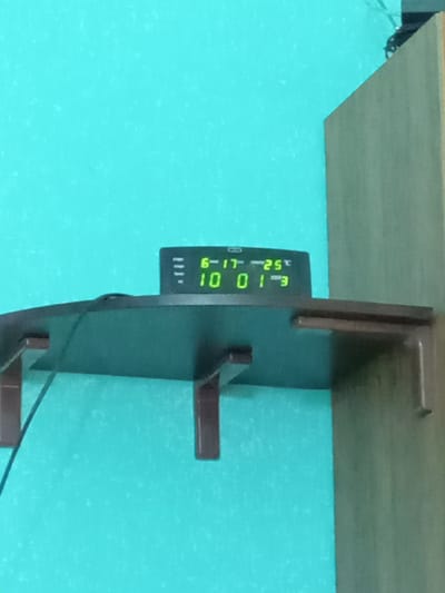 The image shows a close-up of a digital display, likely from a clock or thermometer, mounted on a dark green surface. The display shows green digital numbers and text. Visible text includes "E", "-25°C", "6-17", and "10 01". There are also three small horizontal lines below "6-17". The display is attached to a larger dark object with two horizontal brown shelf-like brackets. In the background, a wooden shelf or surface is visible, with a turquoise wall or surface below it. The image is oriented vertically, with the wooden shelf at the top and the turquoise background taking up most of the frame. The lighting suggests an indoor setting. There are no people visible in the image. The overall scene appears to be a functional display unit mounted in a room. The location context of Kitskany, Moldova is provided but not directly evident from the image itself.