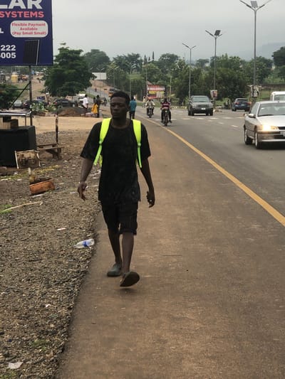The image depicts a man standing on the edge of a paved road in Karu, Nigeria. He is wearing a dark shirt, shorts, and dark shoes, with a yellow safety vest. The road has a yellow line dividing lanes, and vehicles are visible on the road. In the background, there are trees, buildings, streetlights, and signs indicating businesses. A water bottle is visible on the ground near the edge of the road, where the paved surface meets a gravelly area. The scene appears to be a roadside in an urban or semi-urban environment.
