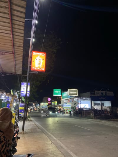The image depicts a street scene at night in Crewek, Indonesia. A person wearing a hijab is seated on a motorcycle in the foreground on the left side of the frame, facing away from the camera. The street is illuminated by various signs and lights from businesses lining the road. Prominently displayed are illuminated signs for "MOMOYO" (advertising ice cream, milk tea, and coffee), "OPPO", and "SAMSUNG". Further down the street, a "VIVO" store is visible, along with signs for "Infinix" and "Farenheit". Several vehicles, including cars and motorcycles, are on the road, some with their headlights on. Pedestrians are also present, walking on the sidewalks and near the storefronts. The environment appears to be a commercial area with shops and food stalls. The overall atmosphere is that of a bustling night market or commercial strip.