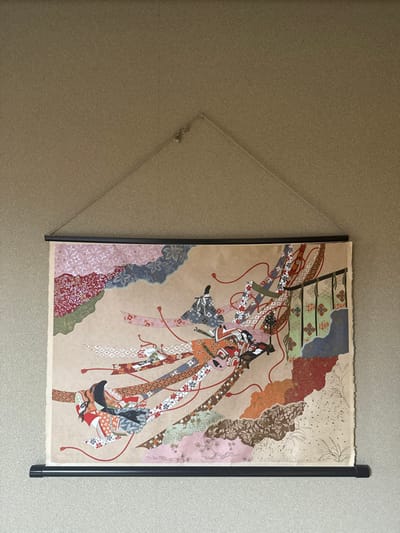 An artwork is centrally mounted on a textured, light brown wall in Nozawaonsen, Japan. The artwork, a print or painting on tan paper with slightly rough edges, depicts three stylized figures in traditional Japanese attire, rendered with intricate patterns resembling collage or fabric applications.
The prominent central figure, positioned diagonally from lower-left to upper-right, is a woman in a multi-colored kimono (orange, red, black, white) with elaborate hair. She appears to be reclining or moving, with numerous patterned fabric strips or streamers extending dynamically from her, intertwined with thin red strings. To her left, she interacts with a small, dark, rectangular object. Above and to her left, a second figure in a gray and black patterned kimono is depicted similarly, with flowing fabric strips and red strings. A third, partially obscured figure, also in a patterned kimono (orange, black, white) with streamers, is visible in the lower-left quadrant.
The artwork's background features abstract, cloud-like, or landscape elements composed of different patterned papers or fabrics in shades of pink, blue, red, green, and brown. On the right side, a stylized architectural element resembling a dark railing or screen with green and orange accents is visible.
The artwork is suspended by a black horizontal bar at the top and another at the bottom, functioning as a scroll hanger. A thin, light-colored cord forms a triangular loop from the top bar to a small, single hook embedded in the wall. A small decorative bell or bead is attached to the cord near the hook. The wall exhibits a faint vertical seam or line extending downwards from the top center.