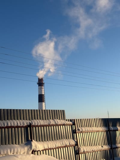 A tall, gray industrial chimney with black and white bands emits white smoke against a clear blue sky with scattered clouds. Power lines stretch across the sky. The chimney is partially obscured by stacked gray corrugated metal panels with rust-colored frames. Patches of snow cling to the frames. The photograph appears to be taken from a low angle, looking upwards.