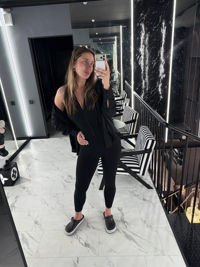 A woman is taking a full-body mirror selfie in what appears to be a modern, upscale retail or commercial space. She is wearing a black, form-fitting jumpsuit with a zipper in the front and black Crocs with white stripes. A black open shirt or jacket is draped over her shoulders. She is wearing a silver cross necklace and a ring on her left hand.
The environment features a black marble-patterned wall with vertical LED strip lighting. To the right, there is a black metal staircase with a black railing and chairs with black and white striped upholstery. The floor is tiled with a white marble pattern. On the left side of the image, there's a mirrored wall reflecting more of the space, and a black cube with a white floral design, possibly a decorative item or a Louis Vuitton inspired piece. The location is the city of Ukromnovske rural settlement, Ukraine.