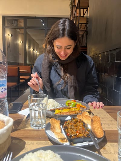 A woman smiles at a table laden with Moroccan food. Skewers of meat and vegetables, a dish of cooked vegetables, and rice are visible on two plates. The table is made of wood with the inscription "LA GRILL." A glass of water and a bottle of water are in the foreground. The background shows a restaurant interior with black-framed windows and chairs. The woman wears a dark jacket and a brown scarf.