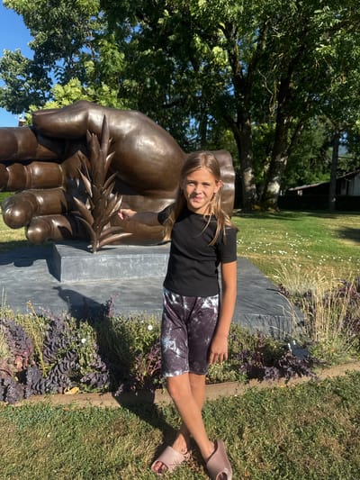A young girl stands in front of a large bronze sculpture of a hand holding a stalk of wheat. The sculpture is located in a grassy area with trees in the background and a small building visible in the distance. The girl is wearing a black t-shirt and patterned shorts, and her right hand is extended towards the sculpture. The scene appears to be outdoors on a sunny day. This sculpture is located at the entrance of the commune of Divonne-les-Bains, France.