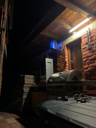 This detailed description pertains to an image captured in Debelt, Bulgaria.

The image depicts an outdoor or semi-enclosed patio area illuminated by artificial light at night. No individuals are present. The primary light source is a long, horizontal fluorescent tube fixture mounted on a brick wall to the upper right. Below it, a large, stainless steel gas grill with a domed lid and control knobs is positioned on a counter. To the left of the grill, a blue light insect zapper is suspended from the wooden ceiling structure, emitting a distinct blue glow.

The wall behind the grill and light fixture is constructed of exposed reddish-orange bricks, and a window with internal blinds or shutters is set into this wall. Various grilling tools, including a spatula and tongs, are hanging on hooks on the brick wall to the right of the window. Several coiled red electrical cables are also visible on the brick wall.

In the foreground, a portion of a dark green or grey outdoor table with a slatted surface is visible. Resting on this table are a coiled black cable, a pair of white sunglasses or safety glasses, and a set of keys on a keyring.

To the far left of the frame, a rough-hewn wooden post or beam is partially visible, from which objects with red handles (possibly knives or utensils) are hanging. In the deeper shadow to the left and mid-ground, a tall stack of dark, rectangular paving stones or tiles is present. The ceiling above is constructed of wooden planks and beams. Strong directional shadows are cast by the artificial lighting, indicating a nighttime environment.