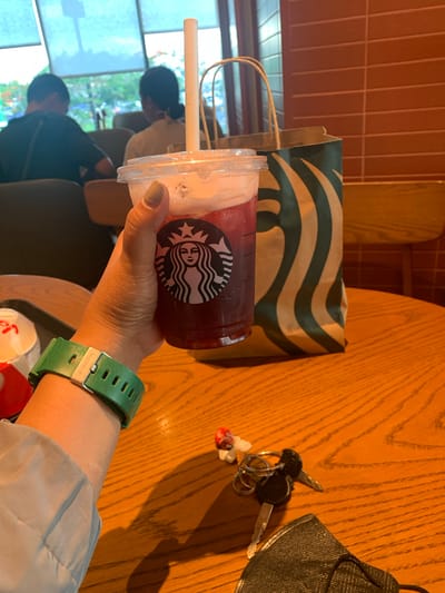 In the city of Angeles, Philippines, a person's hand holds a Starbucks iced beverage with a white paper straw. The person is wearing a green G-Shock watch. A Starbucks paper bag with green stripes is placed on a wooden table. Also on the table are a set of keys, a small red and white decorative item, and a black face mask. In the background, two people are seated near a window with light-colored blinds. The environment appears to be an indoor cafe.