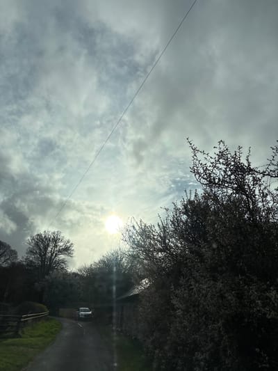 The image shows a partly cloudy sky with the sun shining through. The sun is partially obscured by clouds. In the lower portion of the image is a road flanked by trees and a field. A vehicle is on the road. A power line runs diagonally from the top left. Based on the road configuration, foliage, and vehicle, this scene is possibly located in Shrewsbury, United Kingdom, but without additional landmarks, precise location is difficult to ascertain.