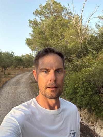 A man with short brown hair and stubble is captured in a selfie. He is wearing a white t-shirt. He appears to be outdoors, with a dirt road curving behind him and lined with trees. The foliage is dense and green, with some bare branches visible. The sky is clear and blue, suggesting it is daytime. The location is a rural area, possibly a hiking trail or a country road. The light suggests it might be late afternoon or early morning, as the sun casts a warm glow. The man's expression is neutral. The setting appears to be in the vicinity of Montuïri, Spain, given the dry, Mediterranean vegetation.