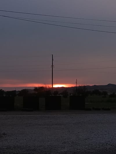 The image depicts a sunset over a rural landscape, likely on the outskirts of Гвардейский, Kazakhstan. The sky is a dusky purple and grey, with a bright orange glow along the horizon where the sun is setting. Several power lines stretch across the sky. In the foreground, there are dark, rectangular structures, possibly concrete barriers, and a gravelly surface. In the middle ground, silhouetted trees and distant hills are visible. A utility pole stands prominently to the right of the frame. The overall impression is one of a desolate, yet beautiful, natural setting.