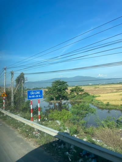The image shows a rural roadside scene in Diên Lạc, Vietnam, under a clear blue sky. A road runs along the left side of the frame, with a metal guardrail separating it from the embankment. The embankment is littered with trash, including plastic bags and bottles, and is covered with sparse vegetation. A road sign with Vietnamese text "CẦU LŨNG Km 1465 + 292 QL.1" is prominently displayed, indicating a bridge and its kilometer marker. Two striped red and white posts stand next to the sign. To the right of the road, a body of water, likely a canal or small river, is visible, with lush green vegetation growing along its banks. Beyond the water, a field of harvested crops, appearing golden brown, stretches towards rolling hills and mountains in the distance. Several trees are scattered throughout the landscape. Overhead, a network of electrical wires crisscrosses the sky. The overall impression is of a sunny day in a Vietnamese countryside setting, with elements of both natural landscape and man-made infrastructure.