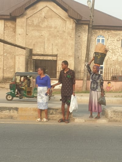 Three people walk along a road's edge next to a building. One woman carries items on her head. A man in a patterned shirt walks between her and another woman in a blue top. A three-wheeled vehicle is nearby. The building behind them is a light-colored, two-story structure with a brown roof, bars on the windows, and a faded mural on an upper wall. The road appears to be asphalt.