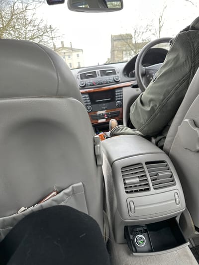 The image depicts the interior of a right-hand drive Mercedes-Benz E-Class (W211 generation) vehicle, viewed from the rear passenger seat looking forward. The grey interior features a central rear console with two air vents and a lower open storage compartment containing a black USB car charger with a green USB-A port and a USB-C port. The driver, wearing a dark green, possibly quilted, jacket, is seated in the front right seat with their hands on the steering wheel. A portion of the driver's leg and a light-colored shoe are visible. The lower leg of a rear passenger, wearing black trousers, is visible in the bottom left foreground. Through the windshield, under an overcast sky, prominent buildings of Dorchester, United Kingdom, are visible: a classical-style, light-colored stone building on the left and the distinctive square stone tower of St Peter's Church, complete with battlements, on the right. Bare tree branches are visible between the car and the buildings. A dashcam or similar device is mounted near the rearview mirror, displaying "1:10".