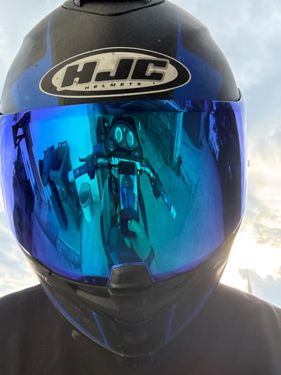 An image, rotated 90 degrees counter-clockwise, depicts a first-person perspective focused on a black and blue motorcycle helmet with a reflective blue visor. The helmet features the "HJC HELMETS" logo prominently on its side.
The highly reflective blue visor captures a complex scene: a person's hand holding a smartphone, seemingly taking a picture. The smartphone's screen, in turn, reflects the helmet again, creating a recursive visual effect. Below the smartphone, the handlebars of a motorcycle are visible, including a grip with "PROTAPER" partially legible, and the motorcycle's dashboard with two circular gauges. Further down in the reflection, what appears to be a somewhat rough or unpaved ground surface is visible, along with a white cylindrical object and a rectangular object.
The background above the helmet shows a bright, partly cloudy sky, indicating that the photo was taken during the daytime. The overall setting suggests an outdoor environment, possibly a rural or unpaved area in Slashten, Bulgaria, viewed from the perspective of someone on or near a motorcycle. The activity is the act of taking a photograph, capturing the helmet, motorcycle elements, and the photographer's device within the visor's reflection.