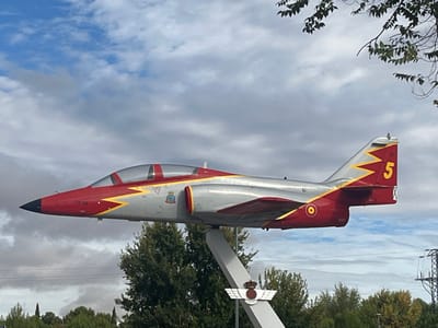 The image displays an outdoor scene in Alcalá de Henares, Spain, featuring a CASA C-101 Aviojet jet trainer aircraft mounted as a permanent monument. This monument is situated at the "Rotonda del Avión" (Airplane Roundabout) on Avenida de Madrid, near the entrance to the Air Logistics Center (CLA). The aircraft is elevated on a light-grey, angled pylon, positioned to simulate flight. Its livery consists of a glossy silver fuselage, red nose cone, tail section, and lower wing surfaces, separated by a yellow zig-zag pattern. Key markings include the serial number "78-08" on the nose, a yellow number "5" prominently displayed on the vertical stabilizer, and the circular Spanish Air Force insignia on the fuselage. A metal crest, depicting a crown surmounting stylized wings, is affixed to the pylon near its base. The background features a partially cloudy sky with patches of blue visible. Dense green foliage from various trees forms the mid-ground and distant landscape, with a leafy branch from a nearby tree partially encroaching into the top right of the frame. Faint utility lines or power poles are visible in the far distance on the right side. No individuals or discernible activities are present in the scene.
