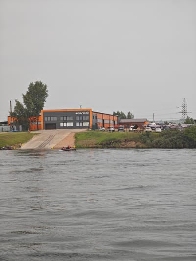 A scene unfolds on the bank of a body of water, likely a river, on an overcast day. A modern, multi-story building with orange and dark grey sections dominates the middle ground. The building features large windows and a prominent entrance with a ramp leading down to the water. The signage on the building, in Cyrillic letters, reads "ФЛАГМАН".
To the left of the building, a tall, slender smokestack rises into the sky, and several trees are present. To the right, a high-voltage power line tower is visible in the distance. The bank of the river is covered with green grass and some low-lying bushes.
In the water, two boats are in motion. One, a dark-colored inflatable boat with several people wearing orange vests, is moving from left to right, creating ripples on the water. Another boat, a red one with people, is further to the left, closer to the bank. On the ramp and the bank, several individuals are observed, some appearing to be near the boats or the building. Several cars are parked on the grassy area next to the building, and a small wooden gazebo is also present. The overall environment suggests a functional area, possibly a marina or a boat service center, located in the city of Peschan, Russia.
