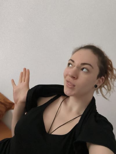 An adult female individual is depicted in a close-up indoor shot against a plain, light grey wall, reportedly located in the city of La Madeleine, France. She has light-colored skin and light brown, wavy hair pulled back. Her attire consists of a black V-neck top with thin straps, covered by an open black garment draped over her shoulders. A thin black cord necklace is visible on her chest. Notable features include a bridge piercing with two silver studs between her eyes and a black circular ear gauge in her right earlobe. Her head is tilted slightly, and her gaze is directed upwards and to the right. Her mouth is slightly open, revealing her tongue partially extended. Her left hand is raised, palm open with fingers slightly spread, positioned to the left of her head. A portion of a brown, possibly fabric, object is visible on the far left side of the frame.