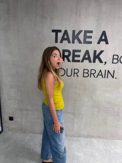 A young woman stands in front of a wall with the words "TAKE A BREAK, BOOST OUR BRAIN" printed on it. She wears a yellow tank top and blue jeans and holds a smartphone. The wall is gray and appears to be made of concrete. The location is likely in an office or public space. The image was taken indoors. It is difficult to verify if the location is Torres Vedras, Portugal from this single image.