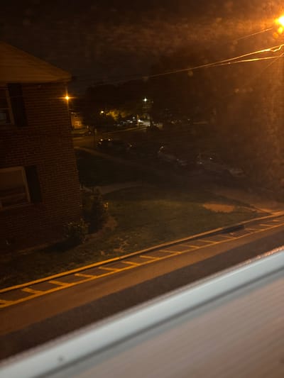 The image captures a nocturnal, elevated view of an urban or suburban area, possibly seen through a rain-streaked window. The composition is diagonal, looking down and across the scene.
On the far right, a broad, light-colored building structure or window frame occupies a significant portion of the foreground, appearing blurred. Adjacent to this, a darker paved surface extends horizontally, featuring clear yellow dashed lines that resemble road or parking lot markings. This paved area runs alongside a strip of dark, indistinct grass or vegetation.
In the midground, the landscape descends into deeper shadows, with a faint, dark shape potentially indicating a parked vehicle. The left side of the image is characterized by several bright, artificial light sources. A prominent, intensely bright orange-yellow streetlight with visible power lines emanates from the upper left, its light diffused and refracted by what appears to be water droplets or smudges on a glass surface. Further down and to the left, another distinct light source illuminates a brick building with visible windows. The periphery of the scene is filled with dense, dark trees and foliage.
The time of day is clearly night, with illumination primarily from artificial streetlights. The atmospheric conditions suggest rain or a wet viewing surface, contributing to the diffused light effects. No people or active events are visible; the scene is static and quiet. There is no visible text in the image. The combination of buildings, streetlights, paved areas, and vegetation points to a developed, inhabited locale.