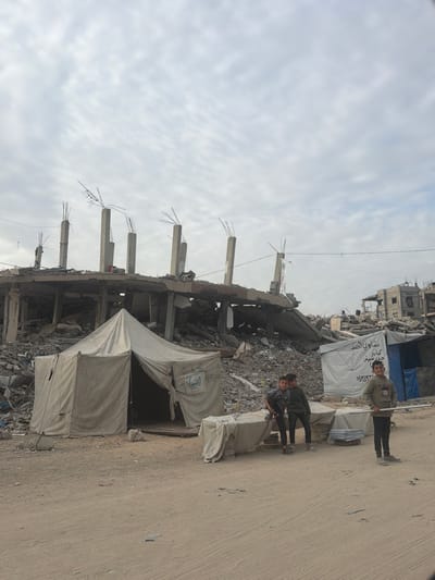 An outdoor scene in Jabalia, Palestinian Territory, depicts an area of extensive structural damage under an overcast sky. In the foreground, an unpaved, light brown dirt or sand surface extends.
In the midground, a light-colored canvas tent, bearing a blue circular logo (likely indicating a humanitarian organization), stands with its entrance partially open. To the right of this tent, three young males are positioned. Two boys stand close together, partially obscured by a low, rectangular object covered with light fabric. A third boy stands further right, facing the camera, holding a light-colored pole horizontally.
The background is dominated by the severely damaged remains of a multi-story concrete building, collapsed into a large pile of rubble. Exposed concrete columns with protruding rebar stand vertically from the debris. Further to the right in the background, additional damaged structures and another light-colored temporary tent, displaying Arabic text and numbers, are visible. The overall environment indicates significant destruction and makeshift living arrangements.
