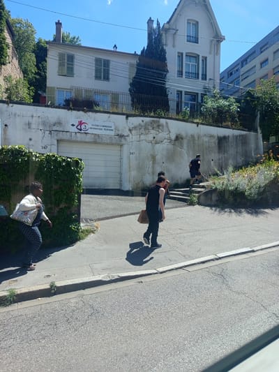 The image shows a street scene in Nancy, France, with several people walking and climbing stairs. In the background, there is a white building with green shutters, a tall building with a blue facade, and a large garage door with a sign that reads "SYSTÈMES D'IMPRESSION INFORMATIQUE - TÉLÉPHONIE". A woman in a patterned dress and white cardigan walks on the left side of the image, carrying a floral tote bag. Several people are walking up a set of concrete stairs on the right side of the image, with one person in a black t-shirt and shorts in the lead. The environment appears to be a sunny day with a clear blue sky.