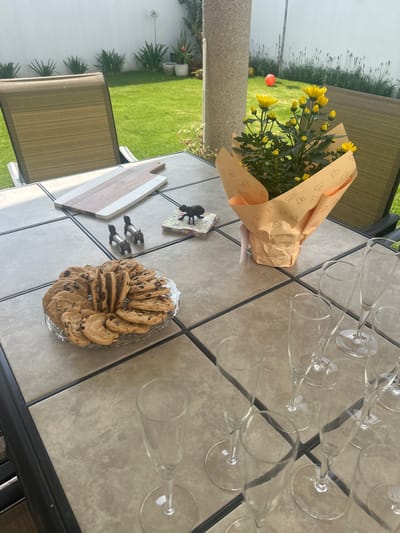 The image shows an outdoor scene, likely a patio or backyard setting in Barrio de Nuevo León, Mexico, given the image's origin. A tiled table is centrally positioned, displaying a still life arrangement of items suggesting a celebratory event or gathering. A bouquet of yellow flowers wrapped in paper with heart designs sits on the table. Nearby, a platter of chocolate chip cookies is displayed. There are also several champagne glasses laid out. Additionally, there is a serving board and decorative objects on the table, and a chair is visible. The background features a green lawn and a white wall, indicating a residential property.
